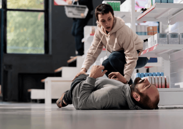 Man lies on the store floor while a woman kneels beside him, offering help near shelving units of products in a retail aisle.