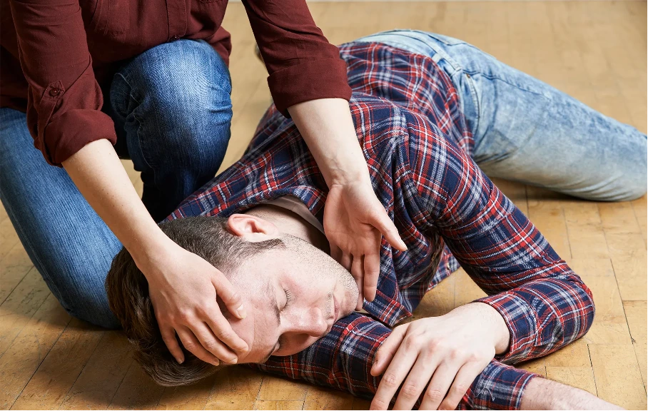 Man lying on a wooden floor in a plaid shirt and jeans while another person supports his head with both hands, suggesting a first-aid or rescue moment.