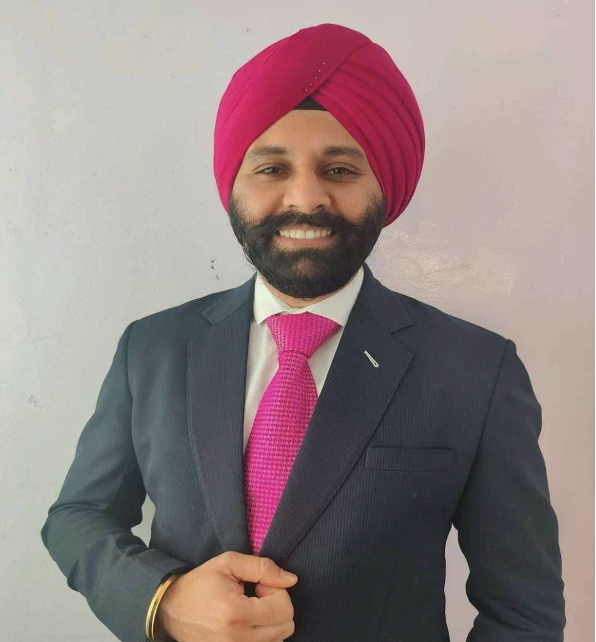 sym Picture 4 | Gurneet Portrait of a man wearing a dark suit and pink turban, smiling at the camera with a full beard.