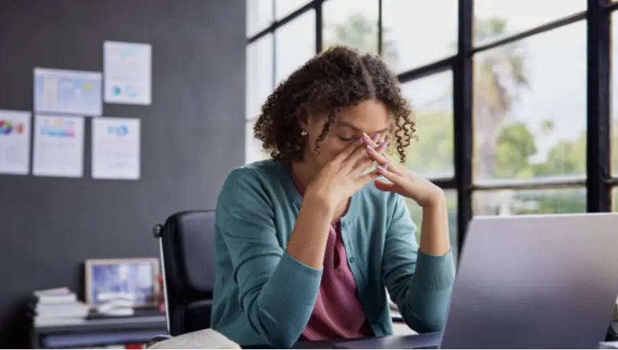 Woman at a desk with hands covering her face, looking stressed while working on a laptop in an office.