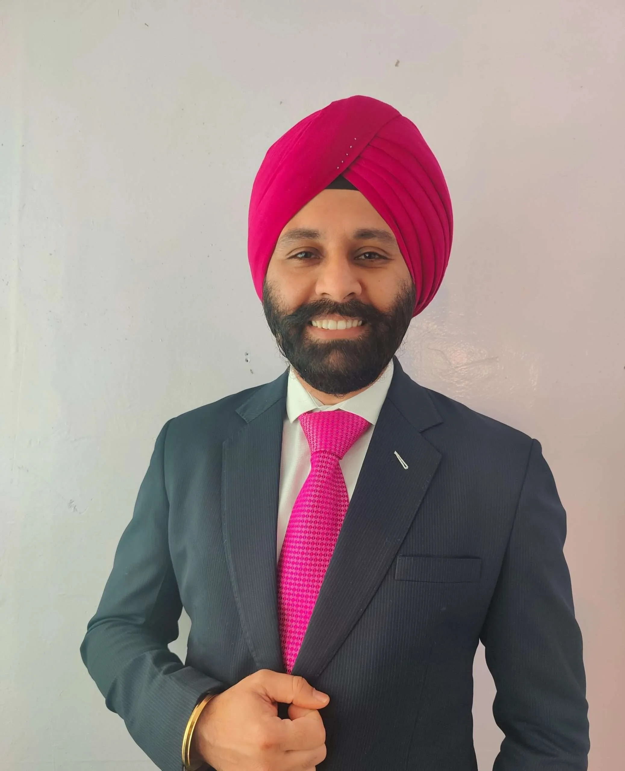 Portrait of a man wearing a bright pink turban and a dark pinstripe suit with a pink tie, smiling at the camera against a plain light background.